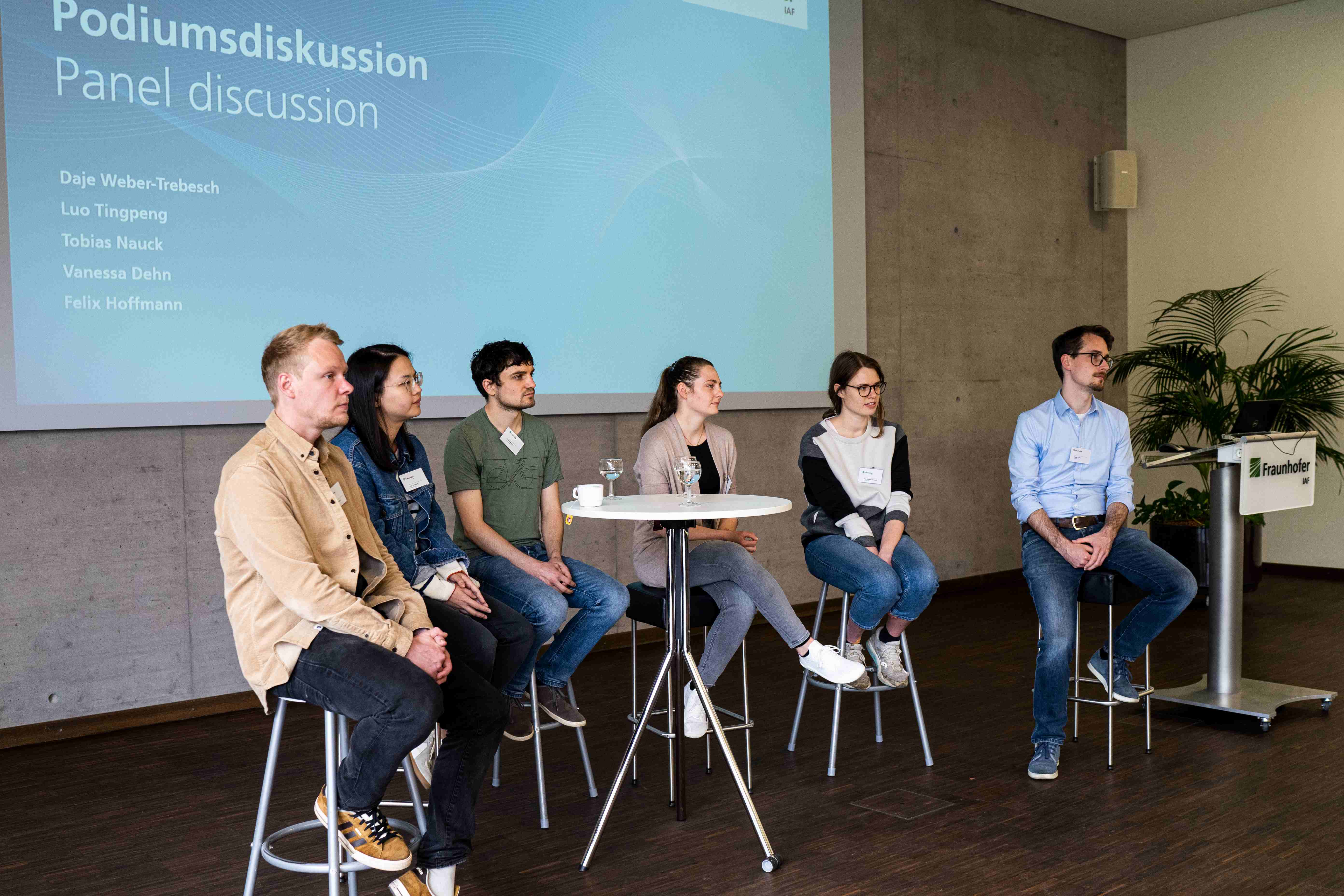 Es sitzen 6 Personen auf Barhocker und hören zu. Hinter ihnen ist eine Folie an der Wand zu sehen, auf der "Podiumsdiskussion" steht.